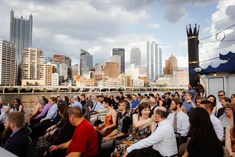 Wedding guests seated on a riverboat's top deck for a ceremony, with the downtown Pittsburgh skyline behind them