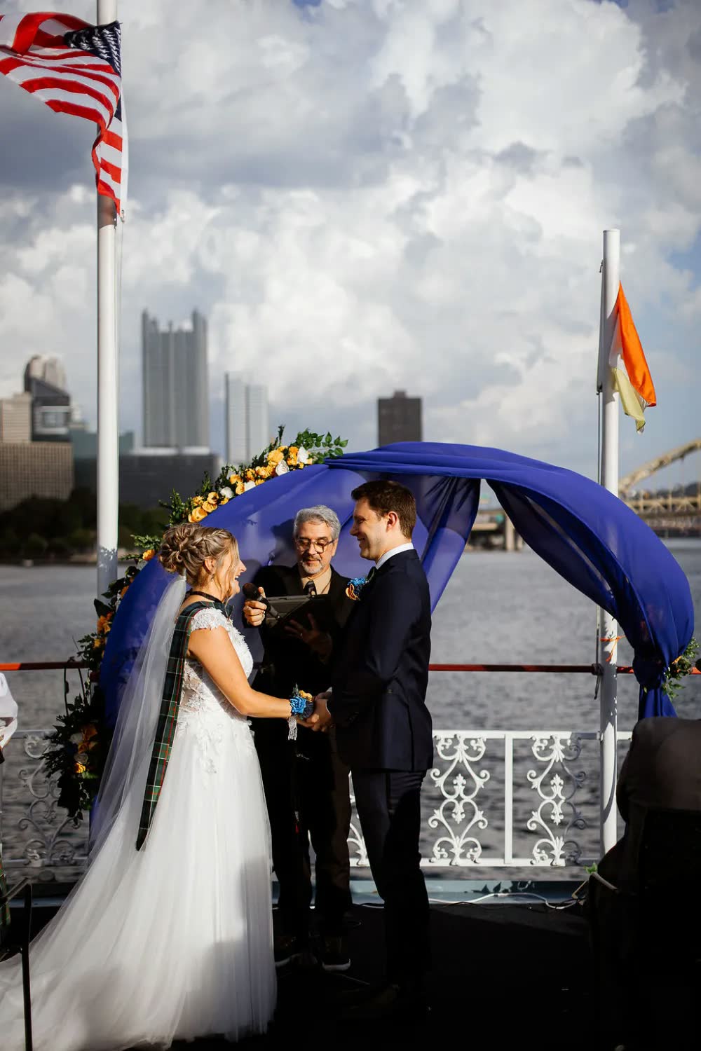 Sheila and Hunter exchange vows under a blue draped floral arch on the top deck as their officiant reads, with the skyline behind them
