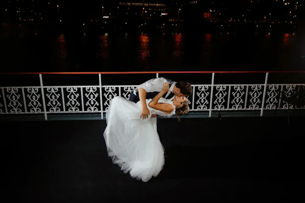 The groom dips the bride for a kiss at the rail at night, the river and city lights behind them