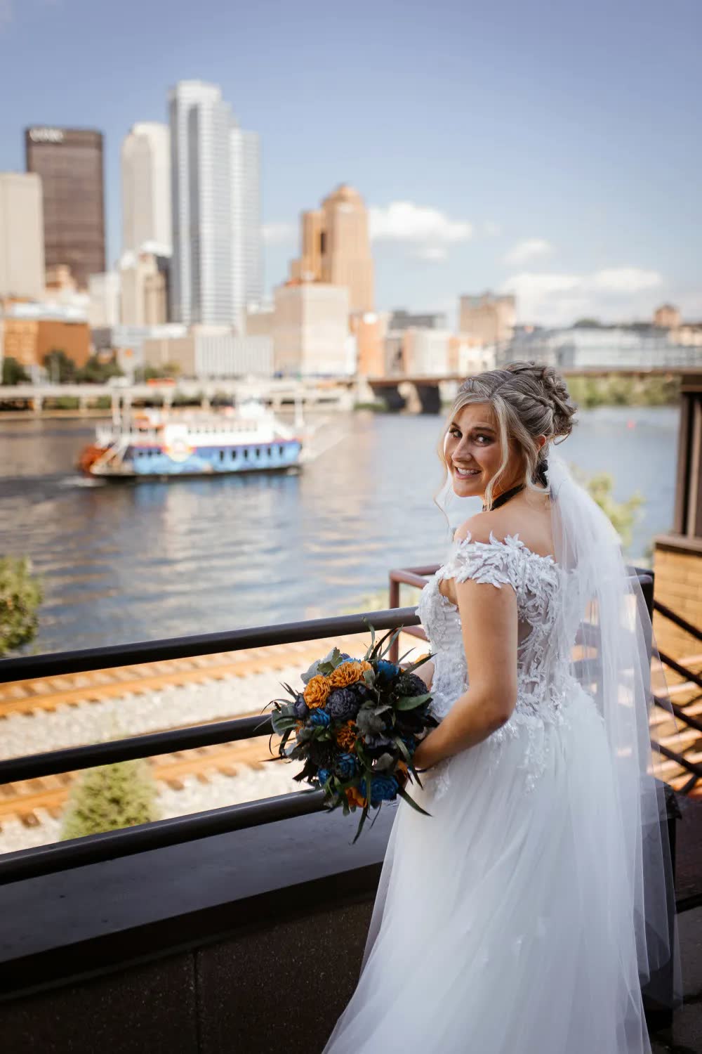 The bride holding her bouquet at the rail, with the Pittsburgh skyline and a Gateway Clipper riverboat on the river behind her
