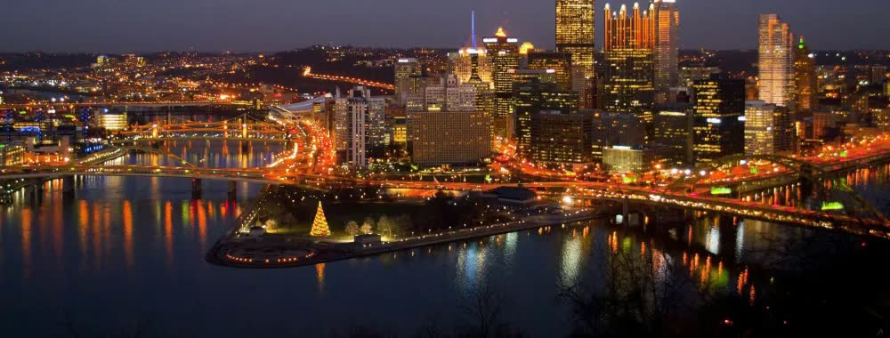 Downtown Pittsburgh lit up at night above the rivers, with a lit holiday tree at Point State Park