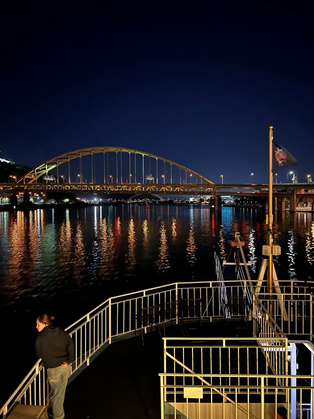 The lit arch of a Pittsburgh bridge reflected in the river at night, seen from the Queen's top deck as a crew member looks out