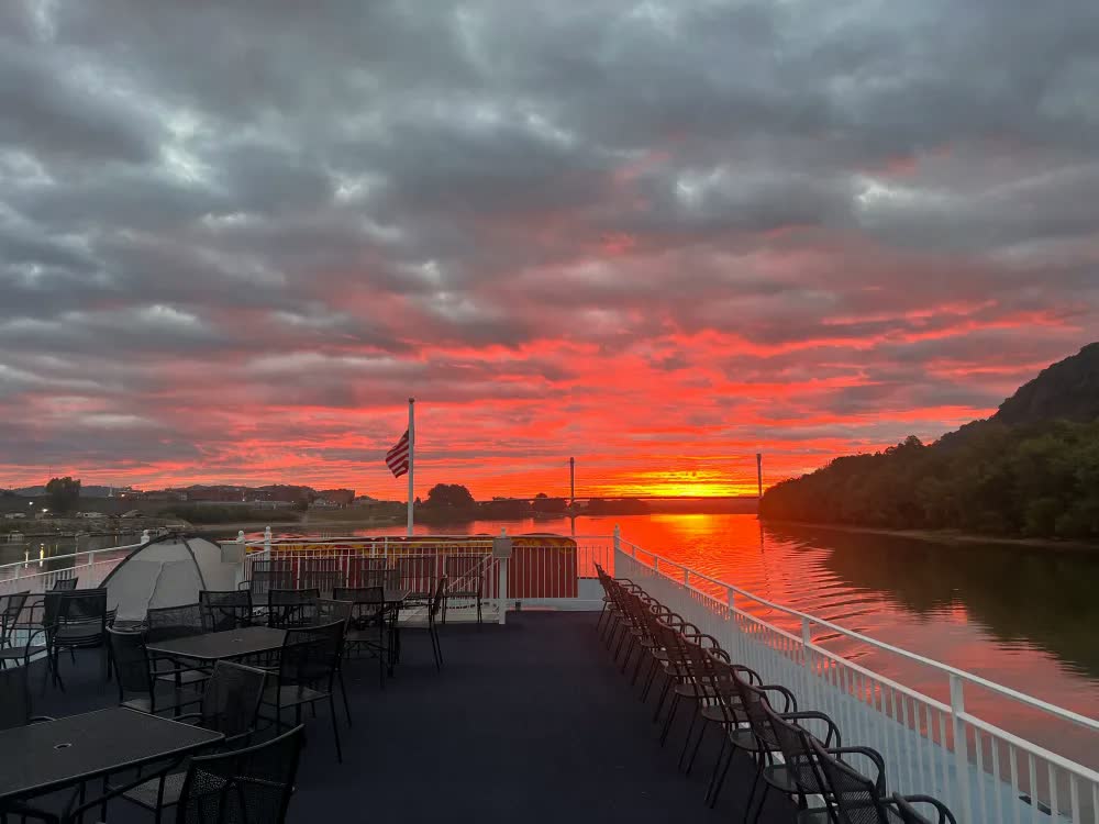 A fiery red sunrise over the Ohio River seen from the empty top deck of the Three Rivers Queen, with an American flag at the bow