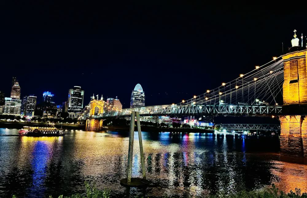 The Cincinnati skyline and the lit Roebling Suspension Bridge reflected in the Ohio River at night, with a riverboat passing