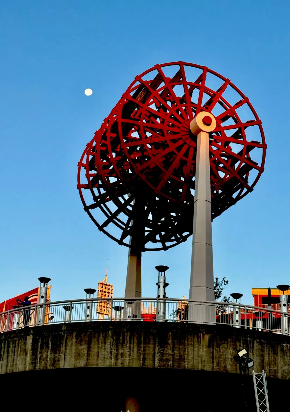A giant red paddlewheel sculpture on tall white pillars against a blue sky, with the moon above