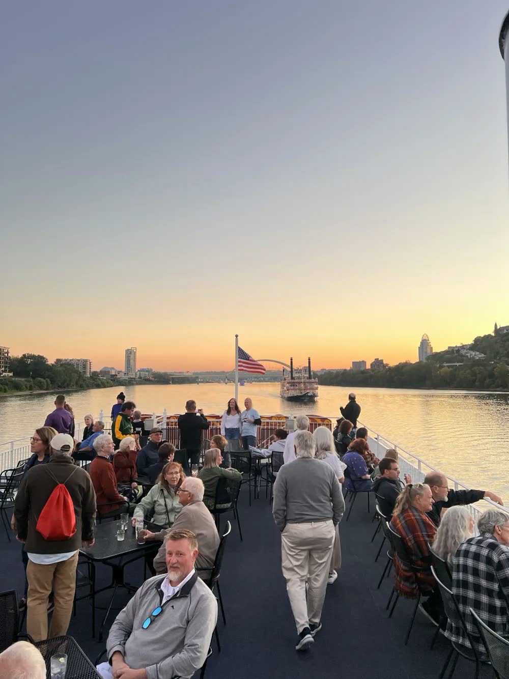 Guests relaxing at tables on the Queen's top deck at sunset, with a paddlewheel riverboat and the Cincinnati skyline ahead
