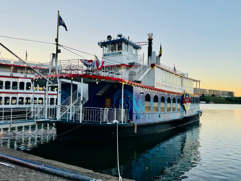 The Three Rivers Queen docked at dusk in Cincinnati, her blue paddlewheel deck and pilot house lit, flags flying