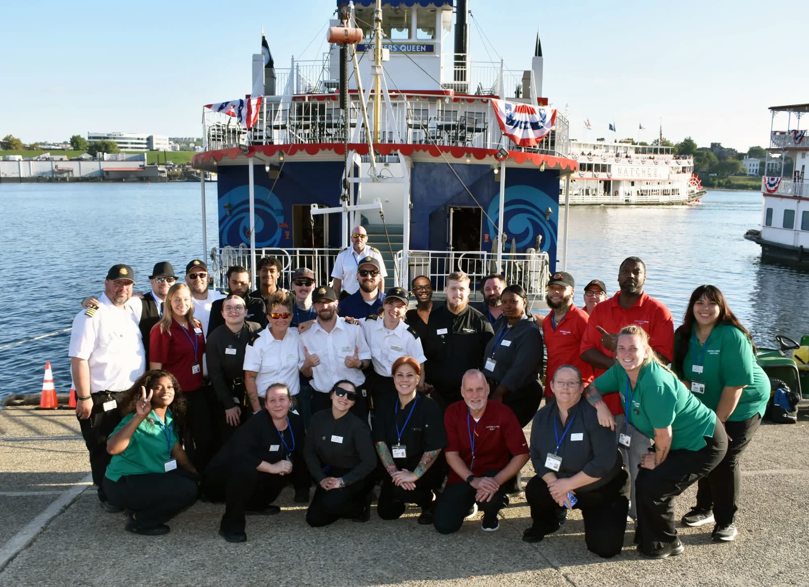 The Gateway Clipper crew in uniform posing on the Cincinnati riverfront in front of the Three Rivers Queen, with the Steamboat Natchez behind