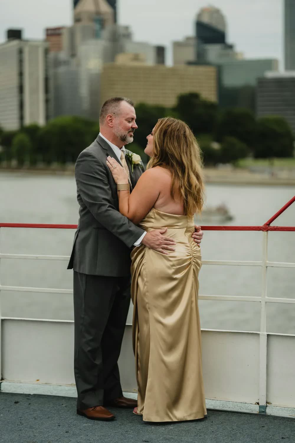 The bride in a gold gown and the groom in a gray suit embrace at the riverboat's rail with the Pittsburgh skyline behind them