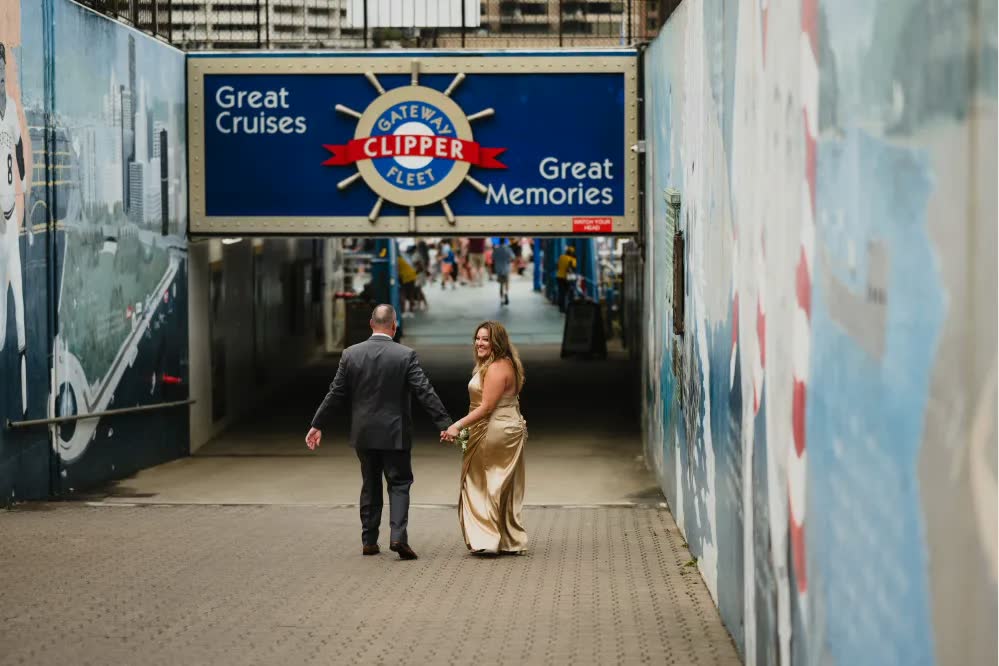 A bride in a gold gown and a groom in a gray suit walk hand in hand through the Gateway Clipper tunnel under the 'Great Cruises, Great Memories' sign