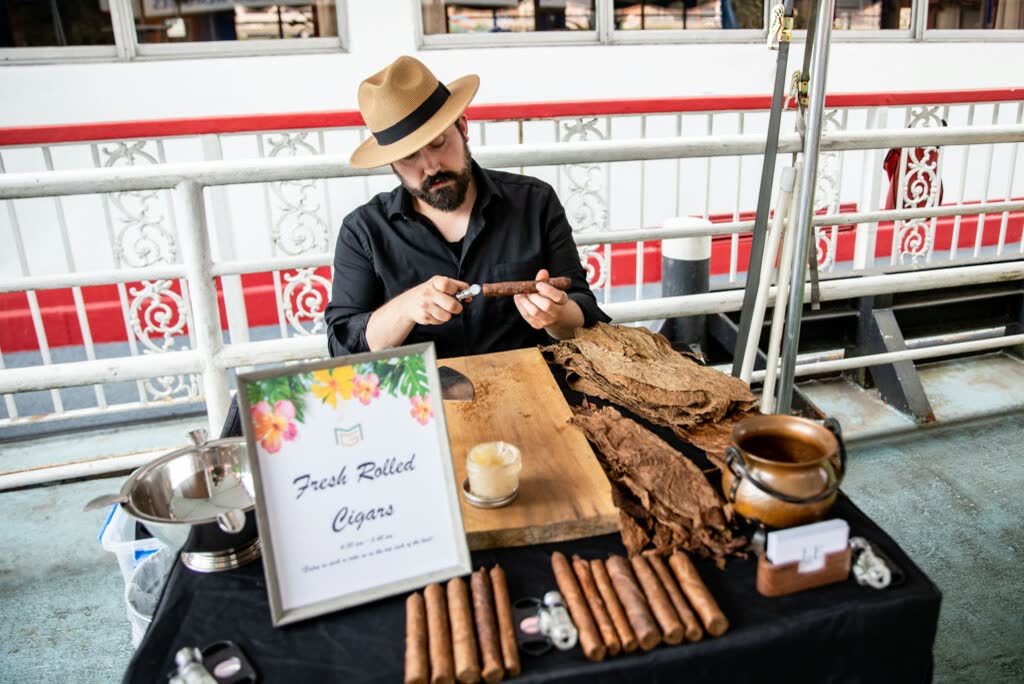 A cigar roller in a straw fedora rolling cigars at a table on the riverboat deck, beside a framed "Fresh Rolled Cigars" sign and a row of finished cigars