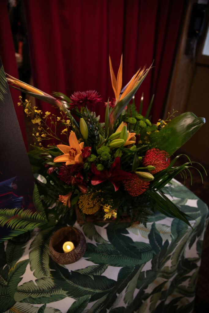 A tropical floral arrangement with bird of paradise, orange lilies and protea on a palm-leaf print tablecloth, with a small candle beside it