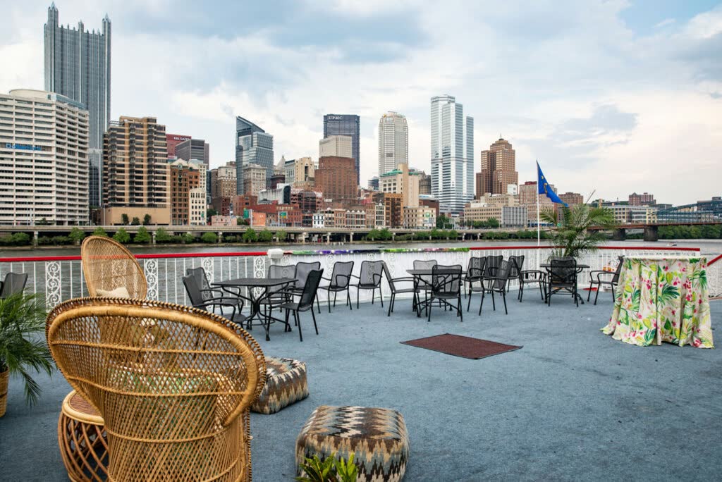 The open top deck of a riverboat set for an event, with wicker peacock chairs, patterned poufs, bistro tables and a tropical-print cocktail table facing the downtown Pittsburgh skyline
