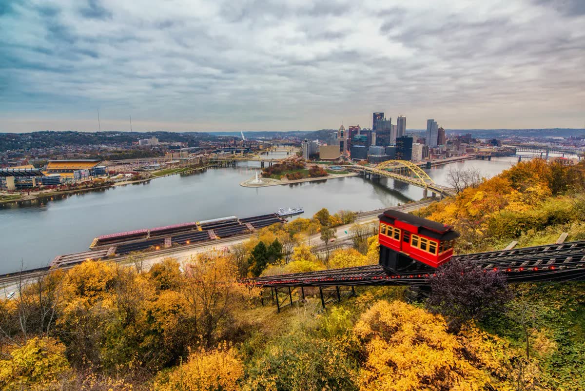 A red Duquesne Incline car climbing Mount Washington through yellow fall foliage above the Point, the three rivers and downtown Pittsburgh
