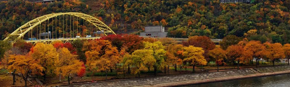 Fall trees in red, orange and gold along the riverbank below a yellow arch bridge in Pittsburgh