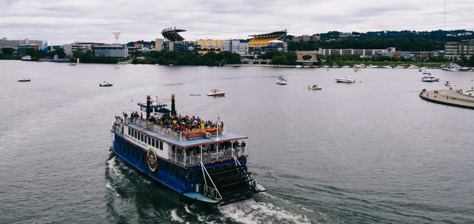 The Three Rivers Queen paddlewheeler, its top deck packed with fans, heading across the river toward Acrisure Stadium