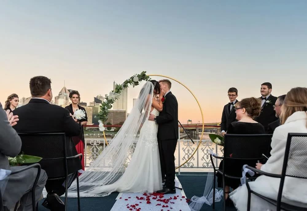 A bride and groom kiss under a round floral arch at a top-deck ceremony at dusk, surrounded by seated guests
