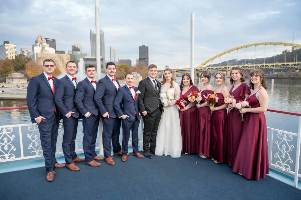 A wedding party in navy suits and burgundy dresses lined up with the bride and groom on a riverboat deck, the Pittsburgh skyline and a yellow bridge behind them
