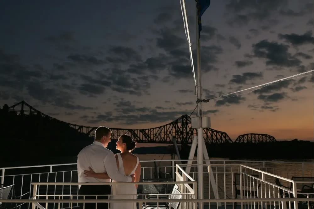 A bride and groom stand together at the rail at dusk, looking out at a river bridge