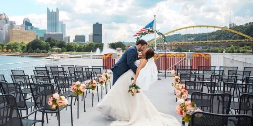 A groom dips his bride for a kiss in the aisle of a top-deck ceremony set with chairs and flowers, with the Pittsburgh skyline and a yellow bridge behind