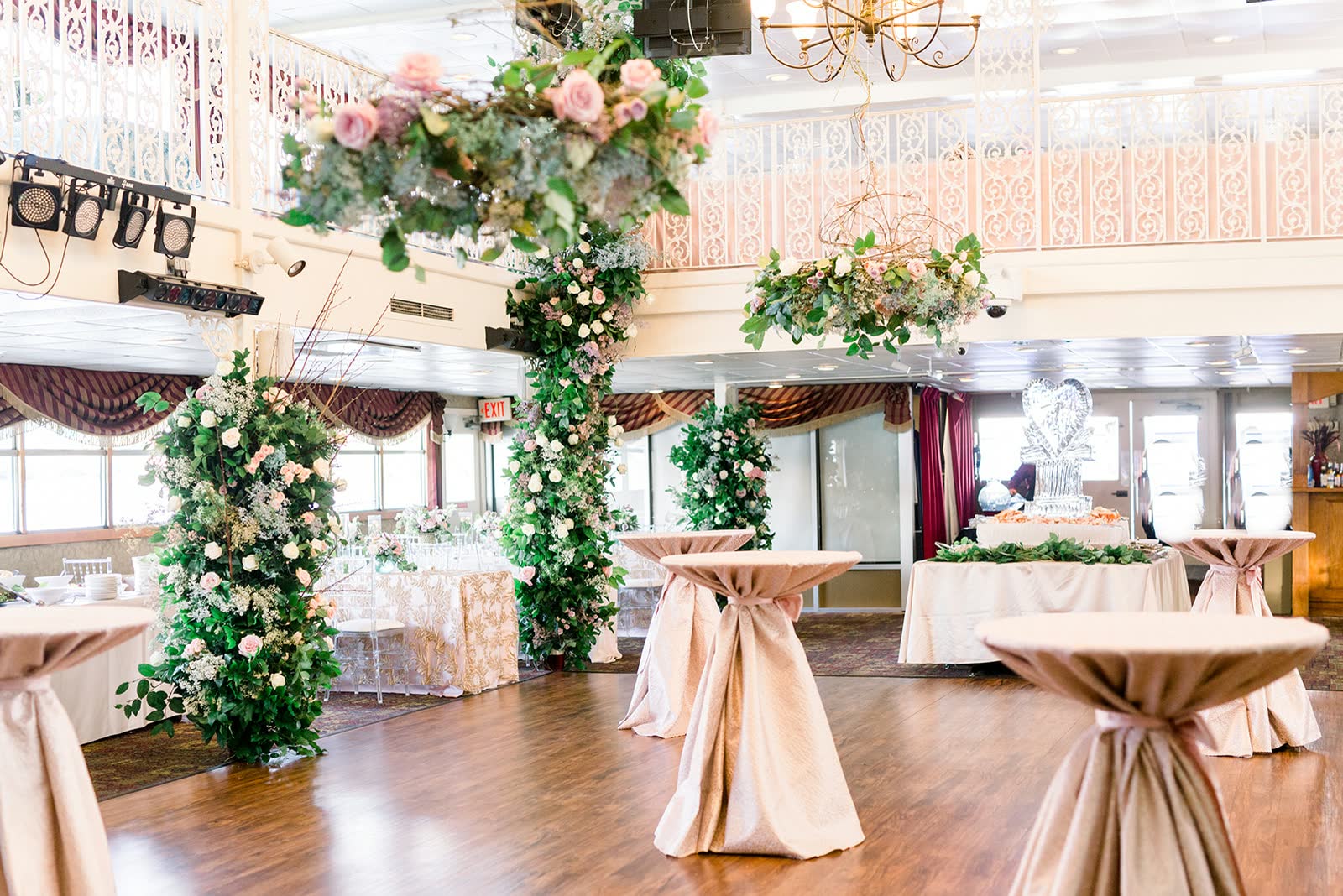A riverboat ballroom set for a reception, with floral columns, cocktail tables draped in blush linen, a wood dance floor and an ice sculpture