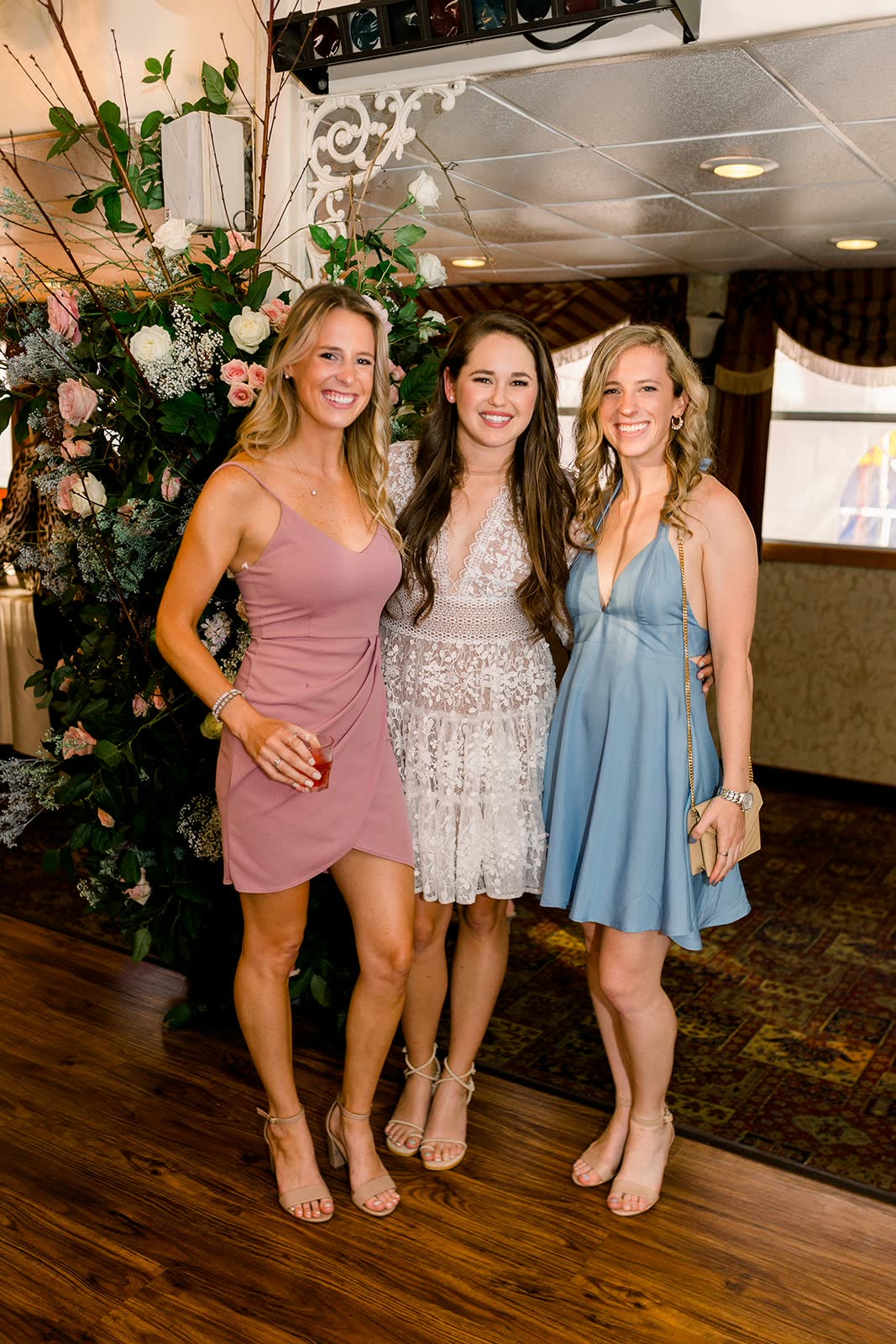 Three friends in summer dresses smiling together in front of a tall floral arrangement inside a decorated riverboat event space