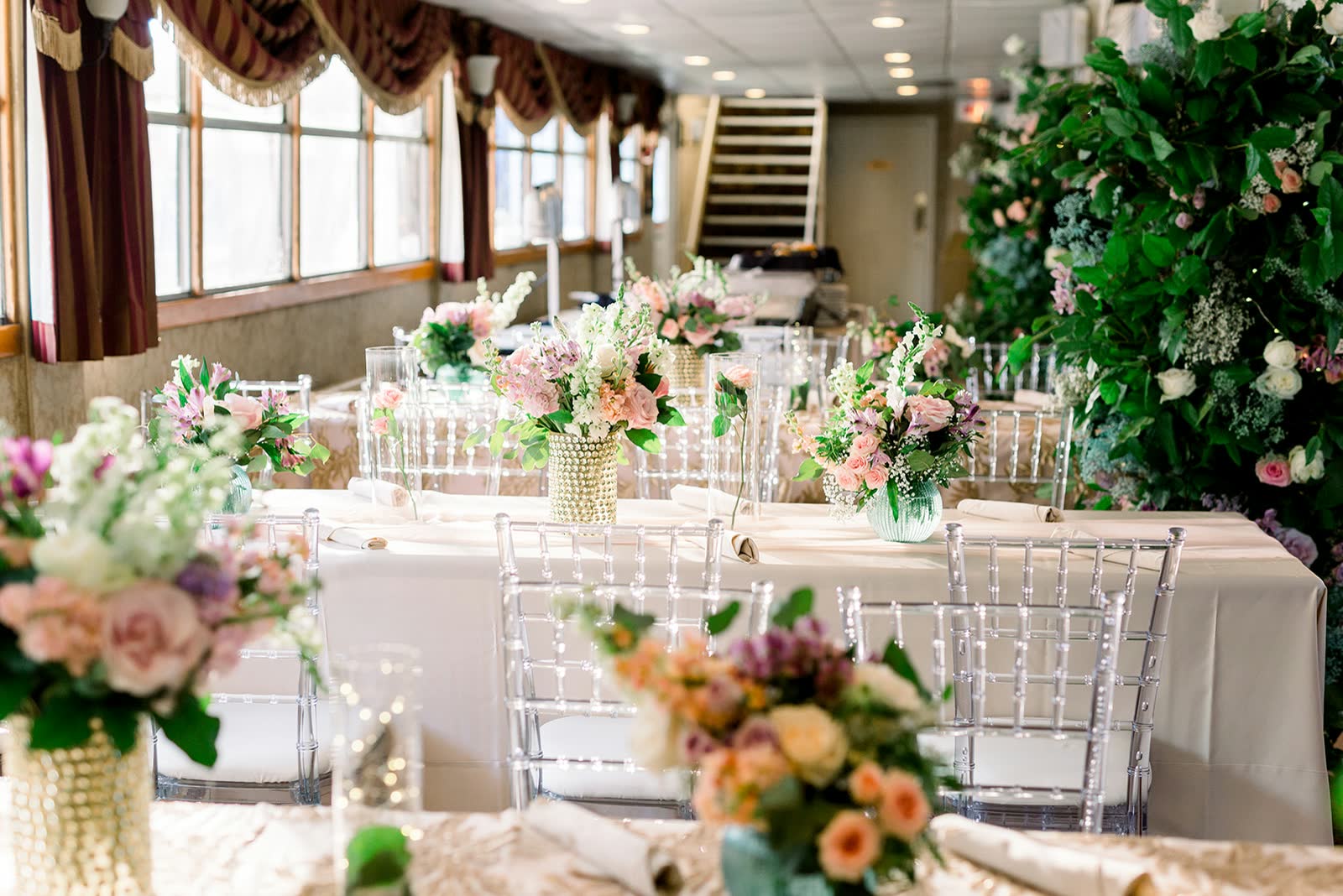 A riverboat dining room set for a wedding, with clear chiavari chairs, white linens and pink and white floral centerpieces beside a wall of greenery and roses
