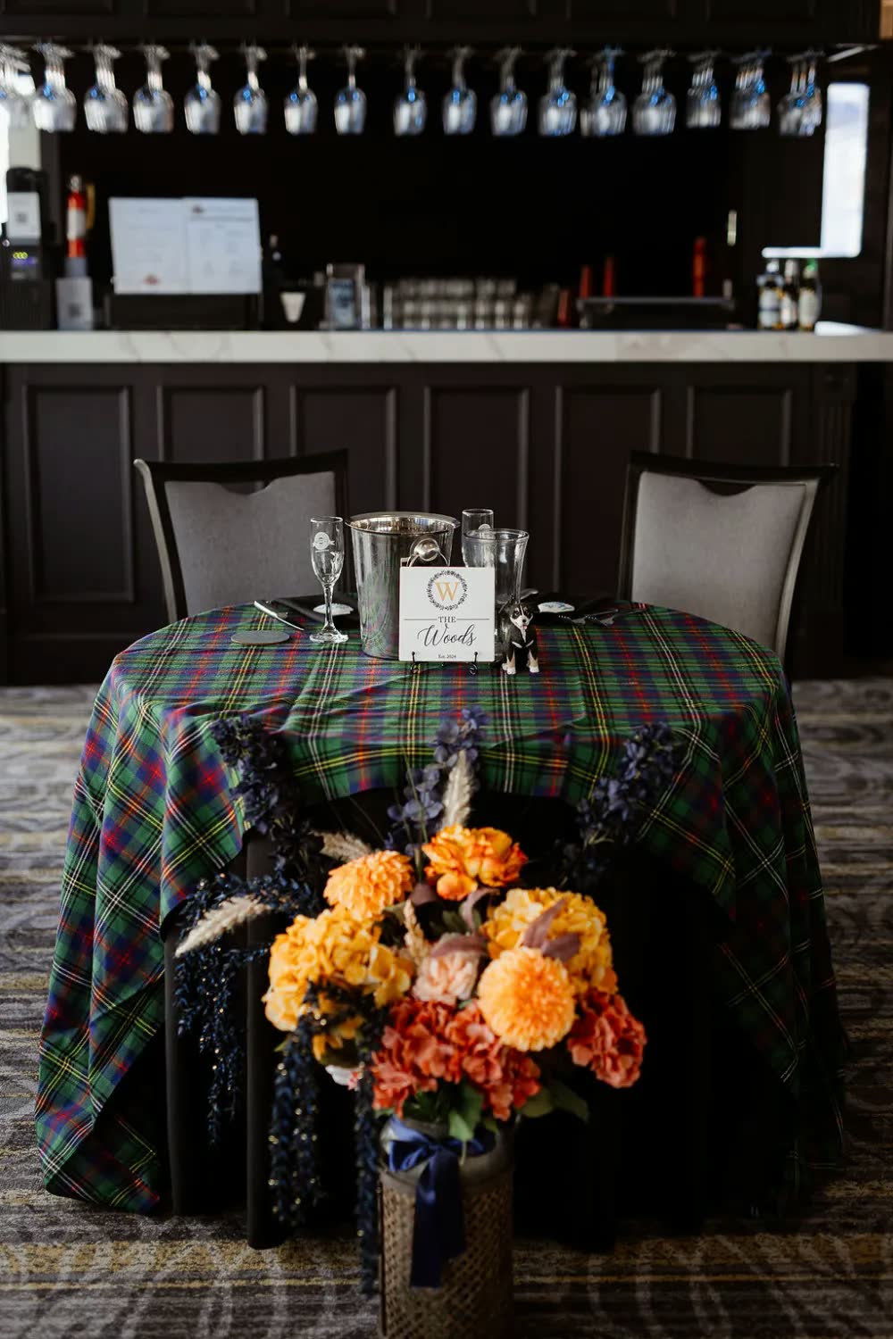 A sweetheart table with a tartan tablecloth and an arrangement of orange and burgundy flowers in front of the riverboat's bar