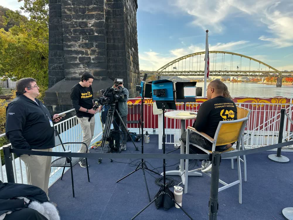A WTAE camera crew and on-air host set up on the top deck of a Gateway Clipper riverboat, with a yellow arch bridge across the river