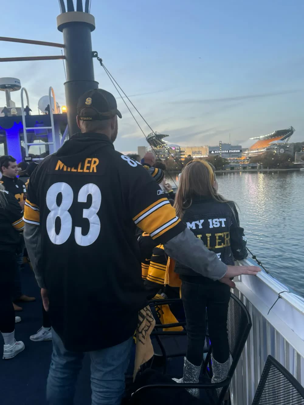 Steelers fans in jerseys on a riverboat deck at dusk, looking across the water at Acrisure Stadium