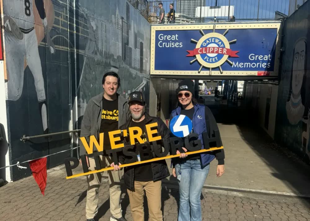 Three people holding a WE'RE PITTSBURGH sign in front of the Gateway Clipper Fleet entrance at Station Square