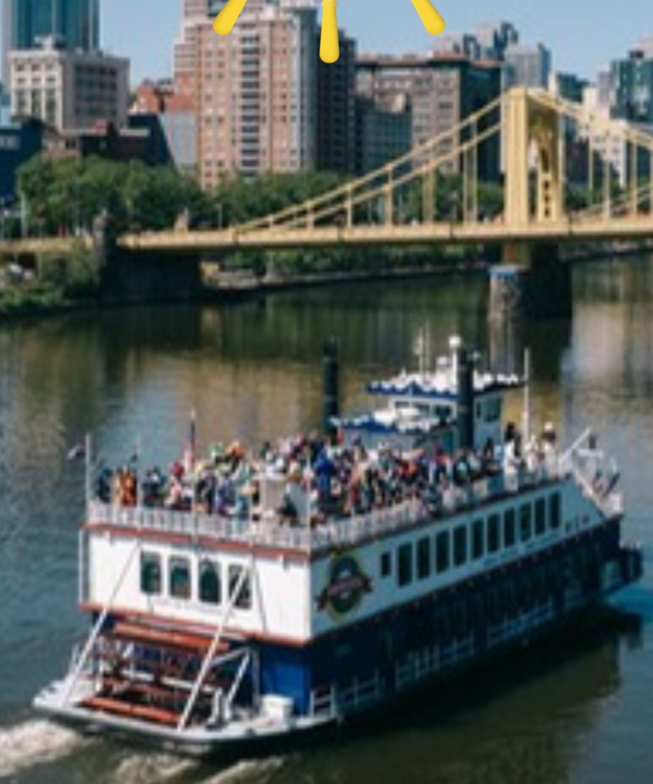 A Gateway Clipper riverboat full of guests on the river below a yellow bridge and downtown Pittsburgh