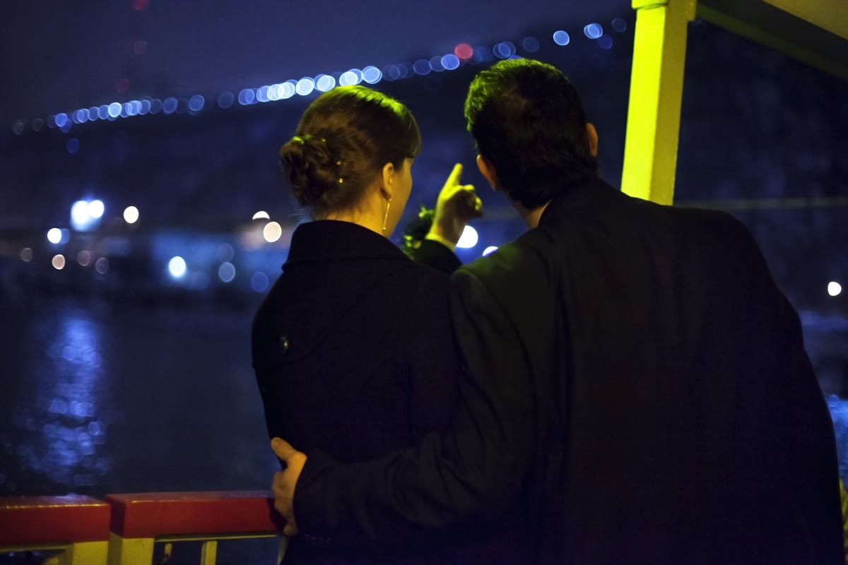 A couple in warm coats at the deck rail of a Gateway Clipper riverboat at night, looking at the lit-up Pittsburgh riverfront