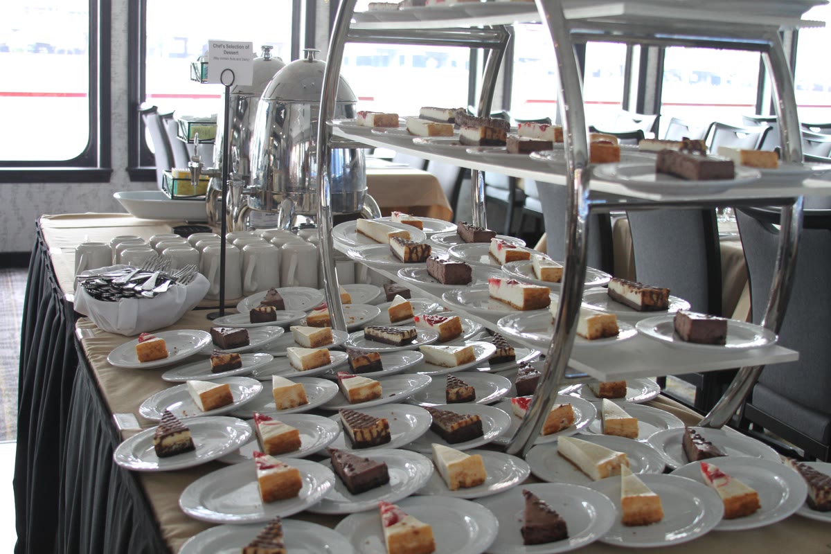 A dessert table of cheesecake slices and chocolate desserts beside coffee urns in a riverboat dining room