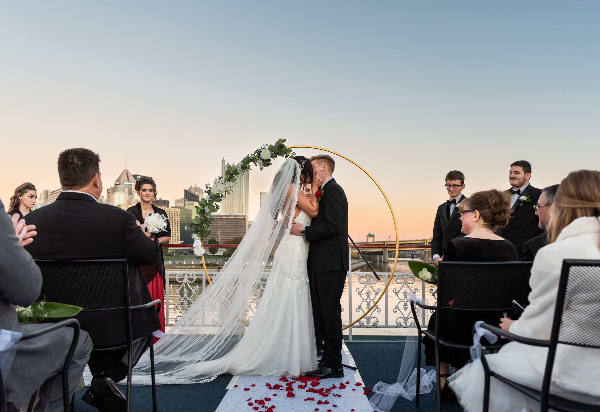 A bride and groom kiss under a round floral arch on a riverboat's top deck at sunset as seated guests look on
