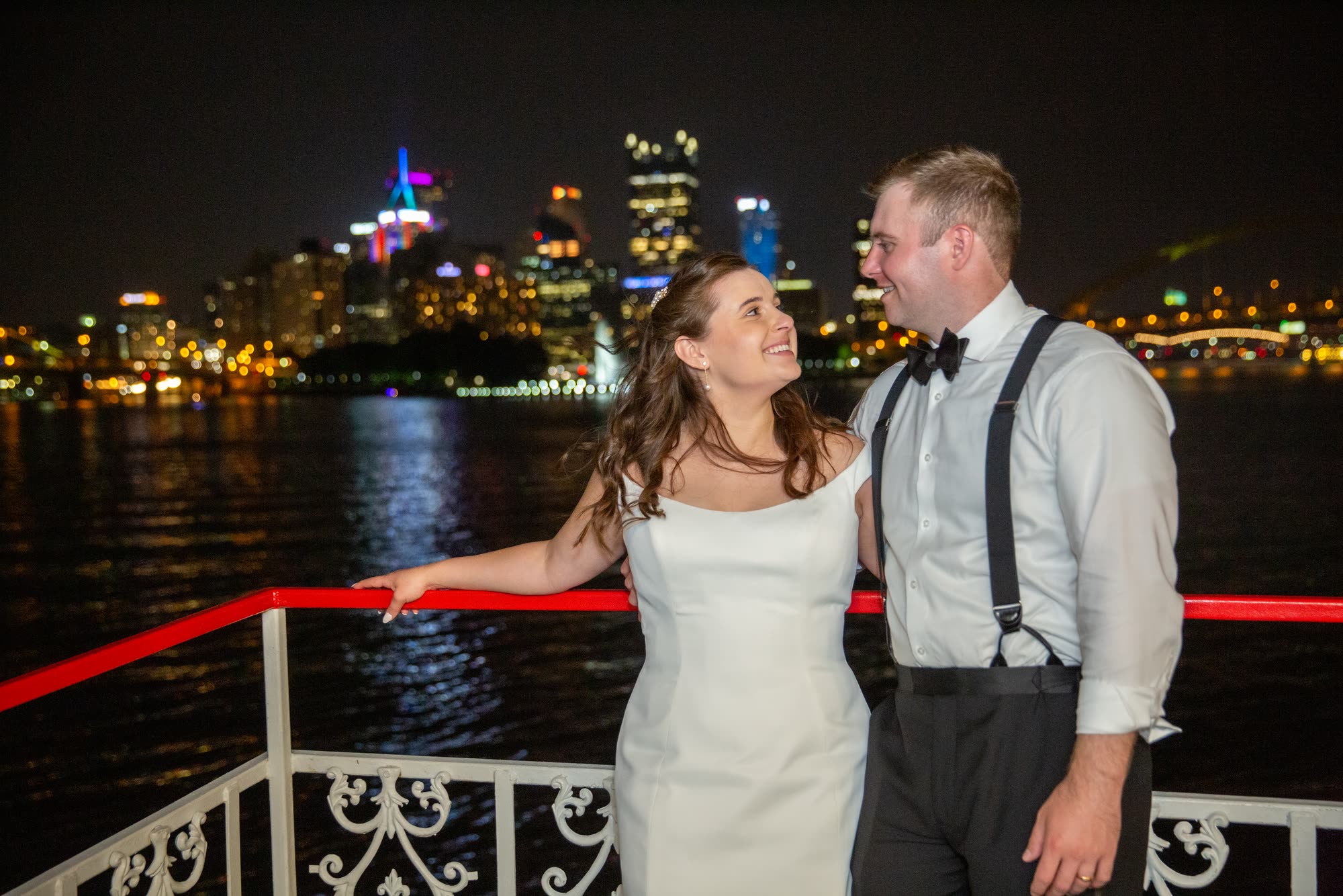 A bride and groom laugh together at the red rail of a riverboat at night with the lit Pittsburgh skyline across the river