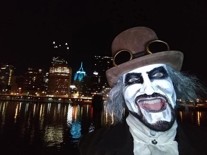 A costumed ghost host in white face paint and a top hat, with the lit skyline behind