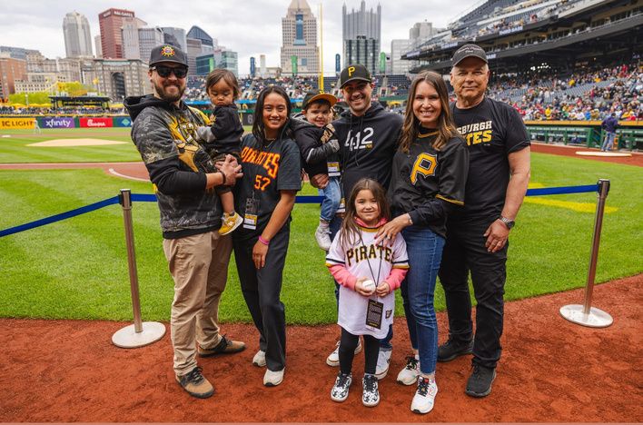 A family in Pirates gear posing on the field at PNC Park, with the downtown skyline behind the stands