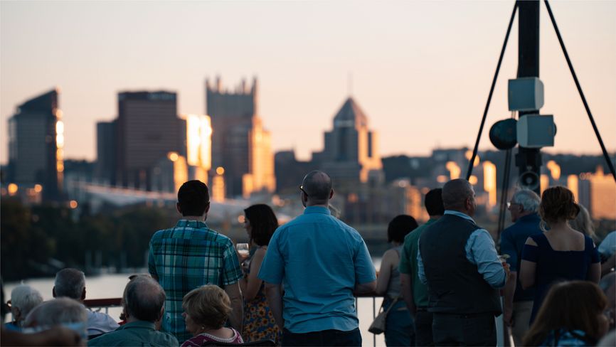 Guests holding drinks on the top deck of a Gateway Clipper riverboat, looking at the Pittsburgh skyline at sunset