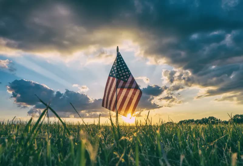 An American flag standing in tall grass against a sunset sky with dramatic clouds