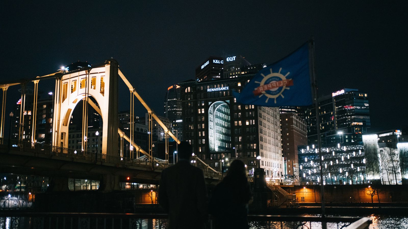 A yellow bridge and downtown lit up at night, with the Gateway Clipper flag