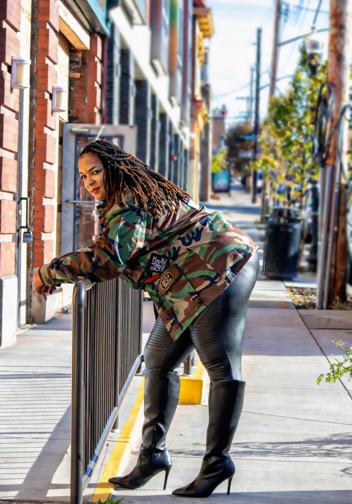 Person in a camo jacket and black boots leans on a rail in an urban setting.