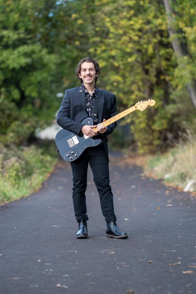 Man in a black suit holding an electric guitar on a forest path.