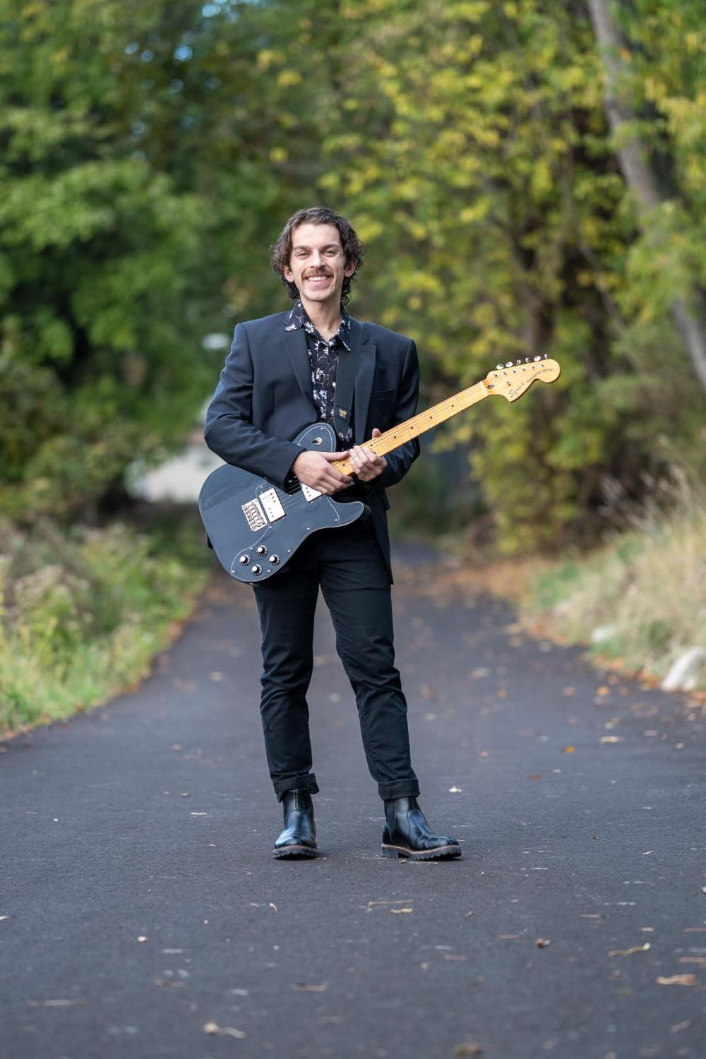 Man in a black suit holding an electric guitar on a forest path.
