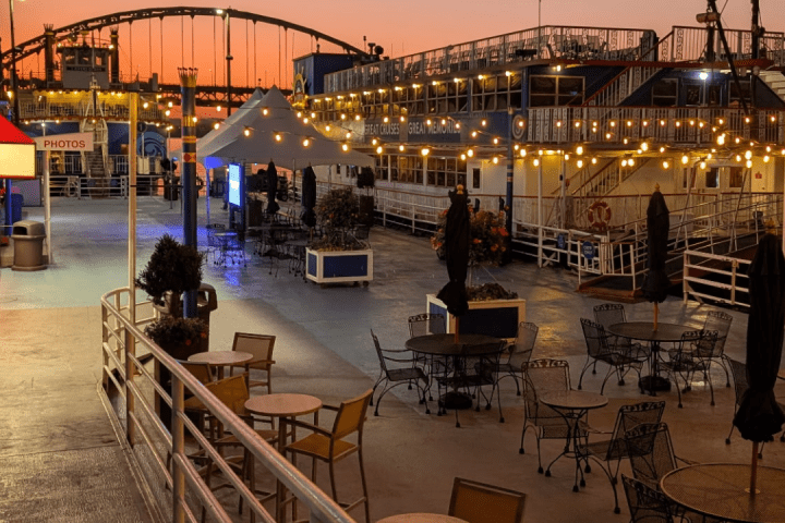 Outdoor seating area with tables, chairs, string lights at sunset beside a docked boat.