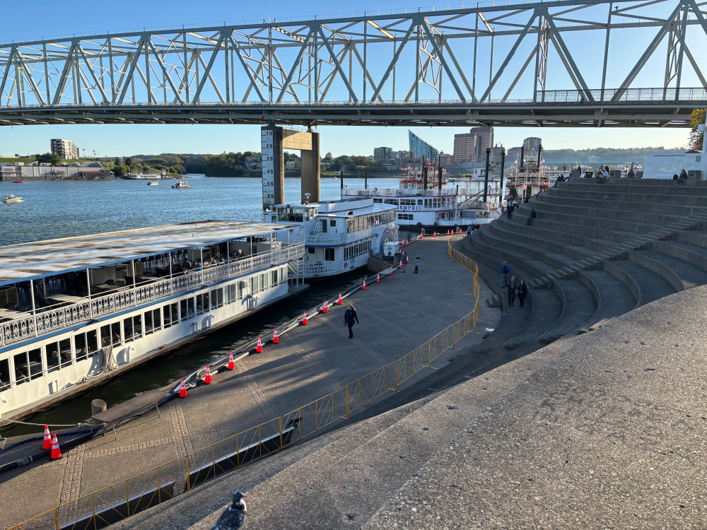 Boats docked by a waterfront under a large bridge with steps and people on the right.