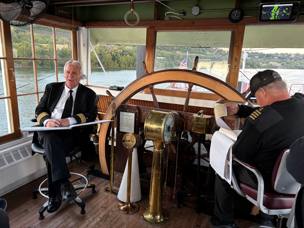 Two men in uniform seated at a ship's wheelhouse with large wooden steering wheel.