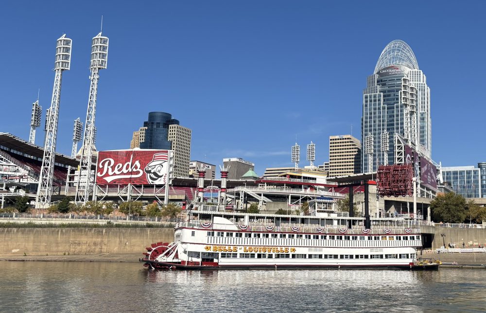Riverboat on water with stadium and city skyline in background on a clear day.