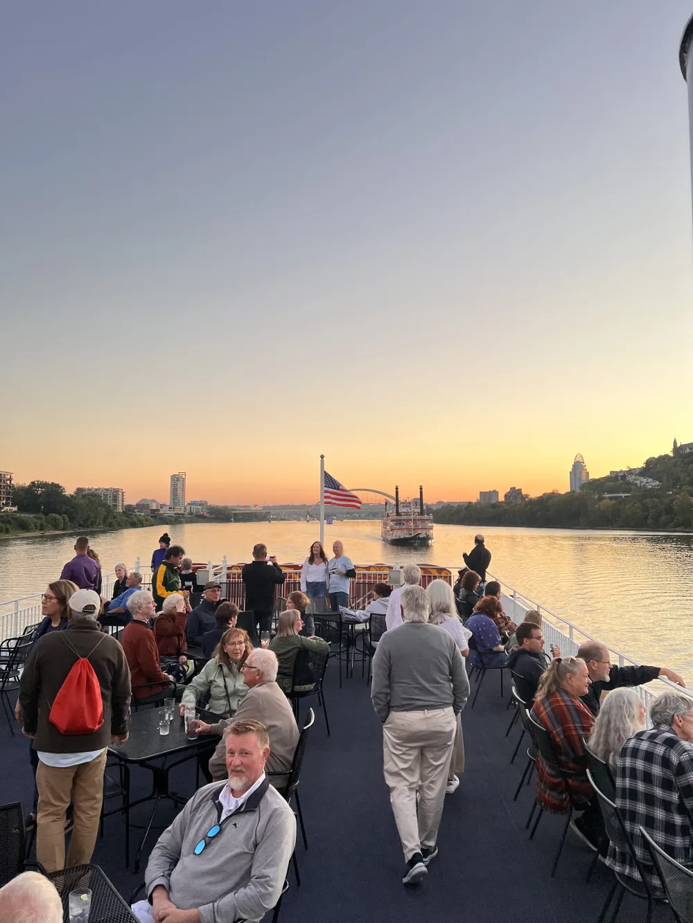 People on a boat deck at sunset, with an American flag and distant city skyline.