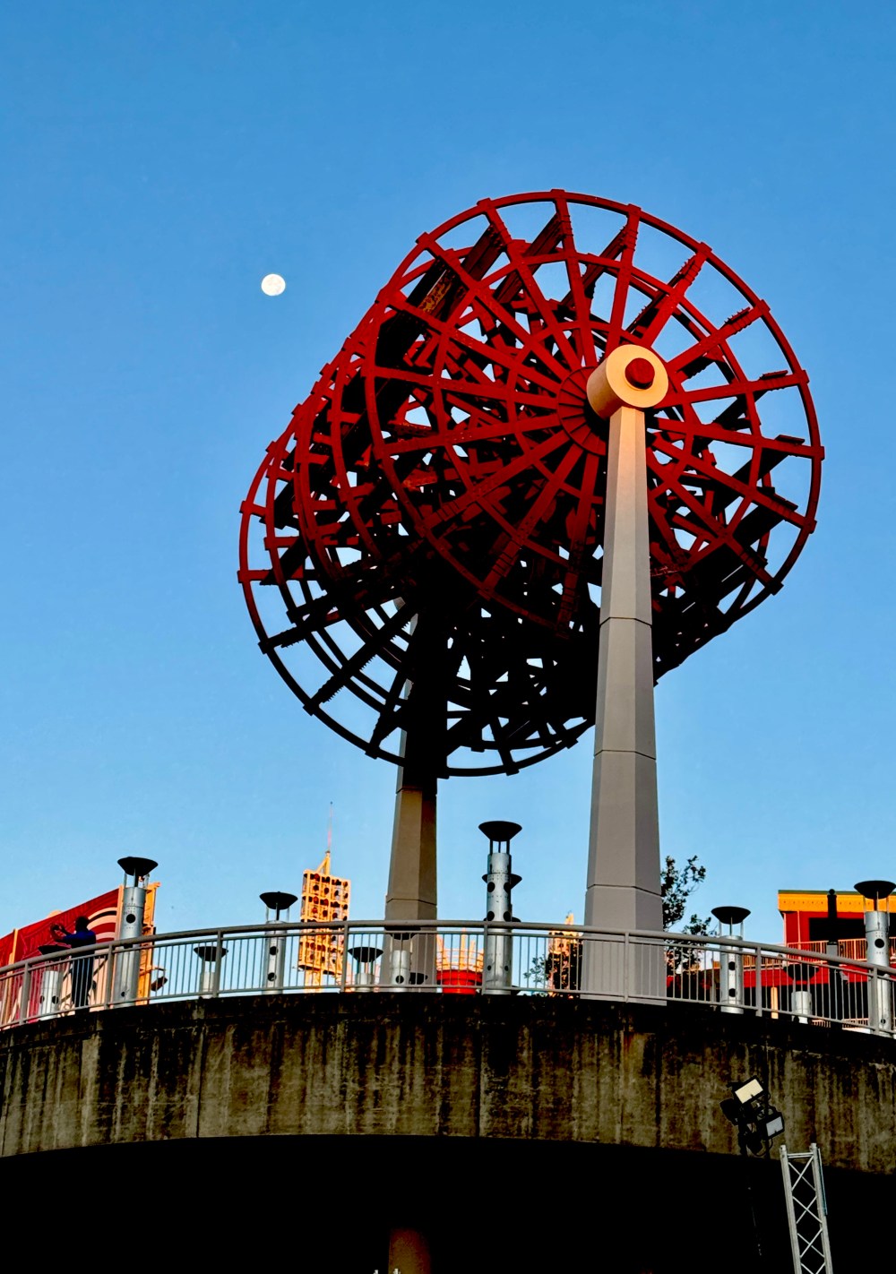 Red sculpture resembling a wheel against a clear blue sky with the moon visible.